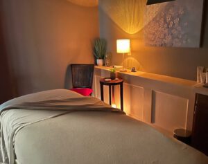 - Results Salon and Spa in Kingsport, TN A massage table with a white sheet in a softly lit room, featuring a lamp, a chair, potted plant, small table, and wall art. - Results Salon and Spa in Kingsport, TN