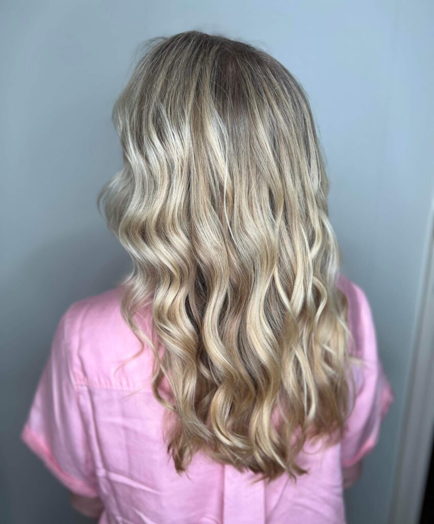 - Results Salon and Spa in Kingsport, TN A person with long, wavy blonde hair wearing a light pink shirt, shown from the back against a plain background. - Results Salon and Spa in Kingsport, TN