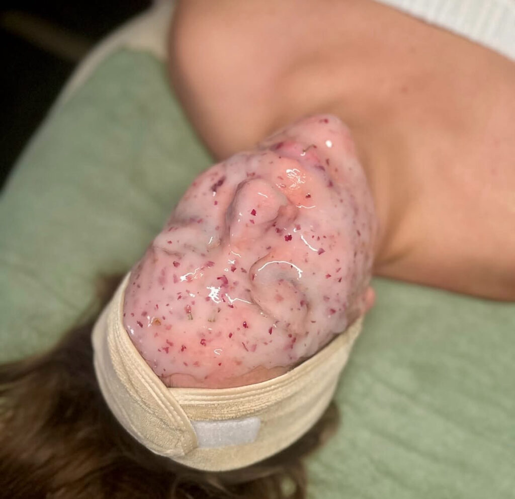 - Results Salon and Spa in Kingsport, TN A person lying down with a pink, textured facial mask covering their face, secured by a beige headband, on a green towel. - Results Salon and Spa in Kingsport, TN