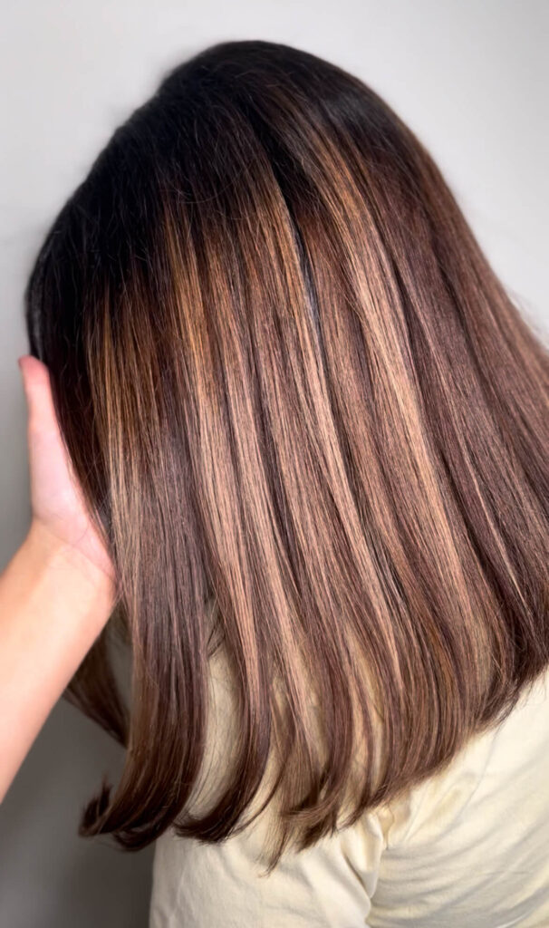 - Results Salon and Spa in Kingsport, TN A person with straight, shoulder-length hair featuring brown and blonde highlights is shown from the back, with a hand touching the side of their head. - Results Salon and Spa in Kingsport, TN