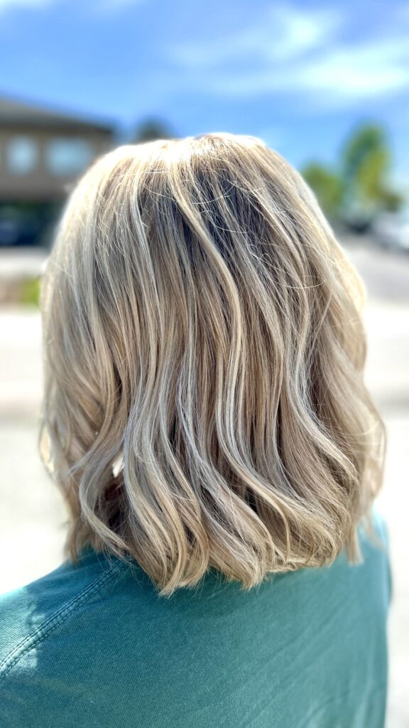 - Results Salon and Spa in Kingsport, TN A person with shoulder-length, wavy blonde hair wearing a green top stands outdoors with a blurred background. - Results Salon and Spa in Kingsport, TN